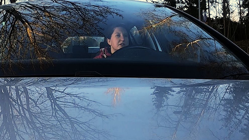 Träd speglar sig i vindruta och motorhuv, i bilen ser man Sofia Holmqvist