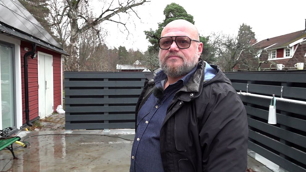 En man med glasögon står på en altan. Bredvid honom syns ett rött hus med ett tak och solpaneler.