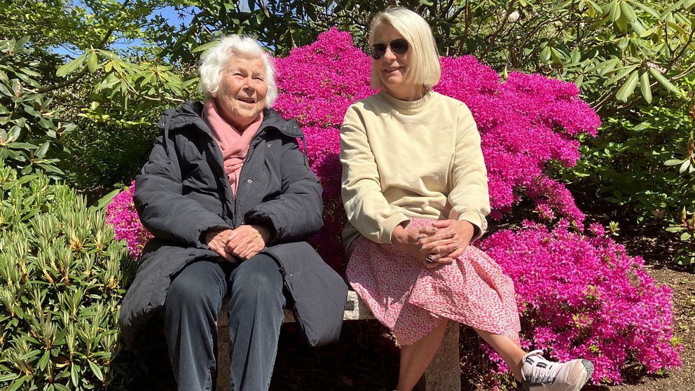 Två kvinnor, en mor och dotterm sitter på en parkbänk. Bakom dem är det fullt av rosa blommor på en buske.