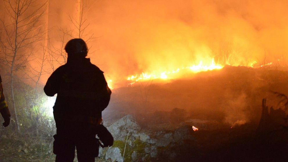 Skogsbrand med öppna lågor och kraftig rökutveckling. I förgrunden syns silhuetten av en person från räddningstjänsten.