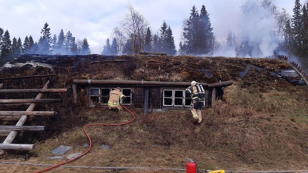 Två brandmän släcker en brand i Jormvattnet. Byggnaden är välvd med torvbeklätt tak. Rök syns stiga från byggnaden, men inga synliga lågor.