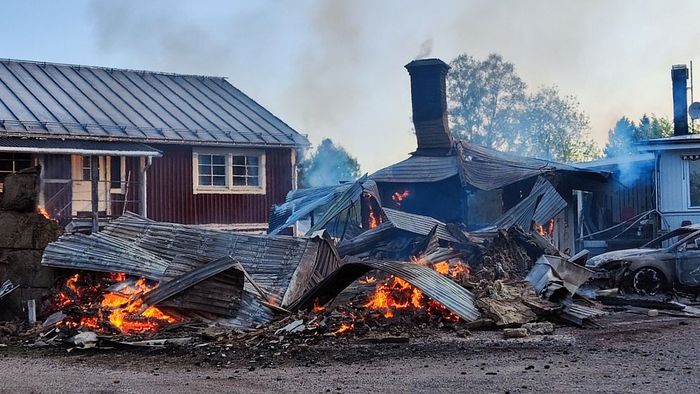 På bilden syns en byggnad som brunnit ned till grunden. På bilden syns fortfarande eldlågor och ovanför ligger plåttak.