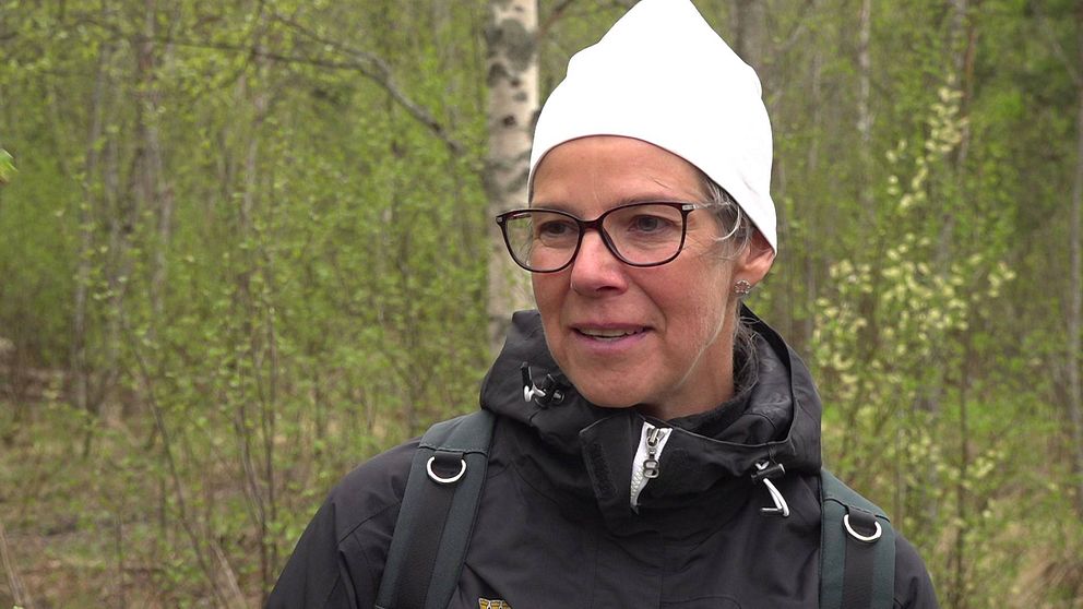 Kvinna med vit mössa, svart jacka och svartbågade glasögon står utomhus med grönskande skog bakom sig.