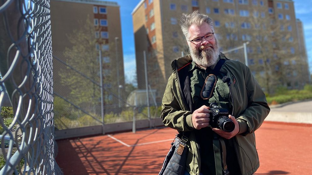 Man med skägg håller i kamera på en idrottsplan framför höghus i miljonprojektområde.