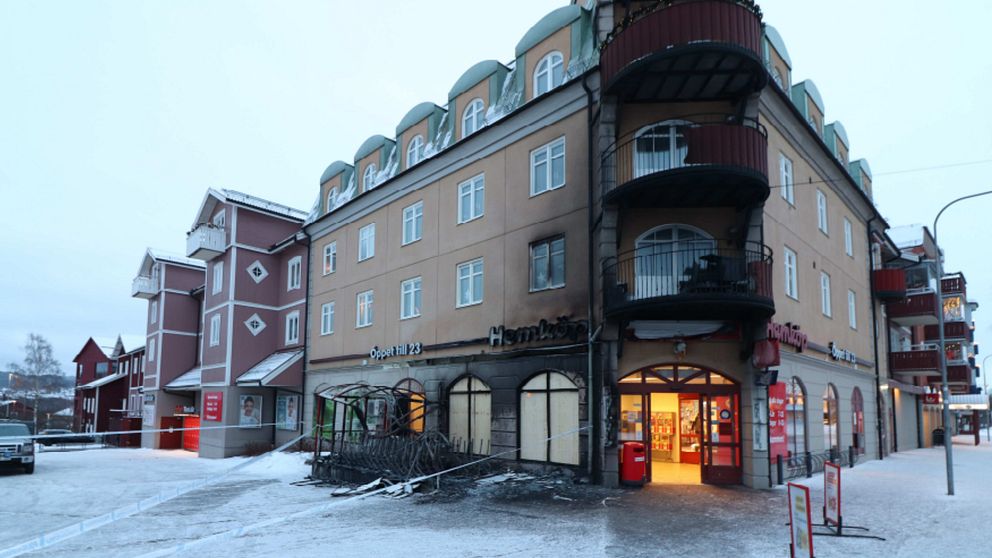 Mörk sot ligger över tre balkonger – nedanför ser man att entrén till matvaruaffären är alldeles svidd och att utrymmet där kundvagnar parkeras är nedbränt.