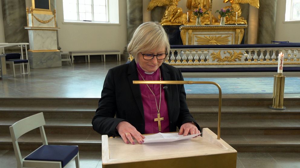 En bild på en kvinna, biskopen Susanne Rappmann, i rosalila skjorta, prästkrage, kavaj och korshalsband står vid en predikstol med ett papper i handen.