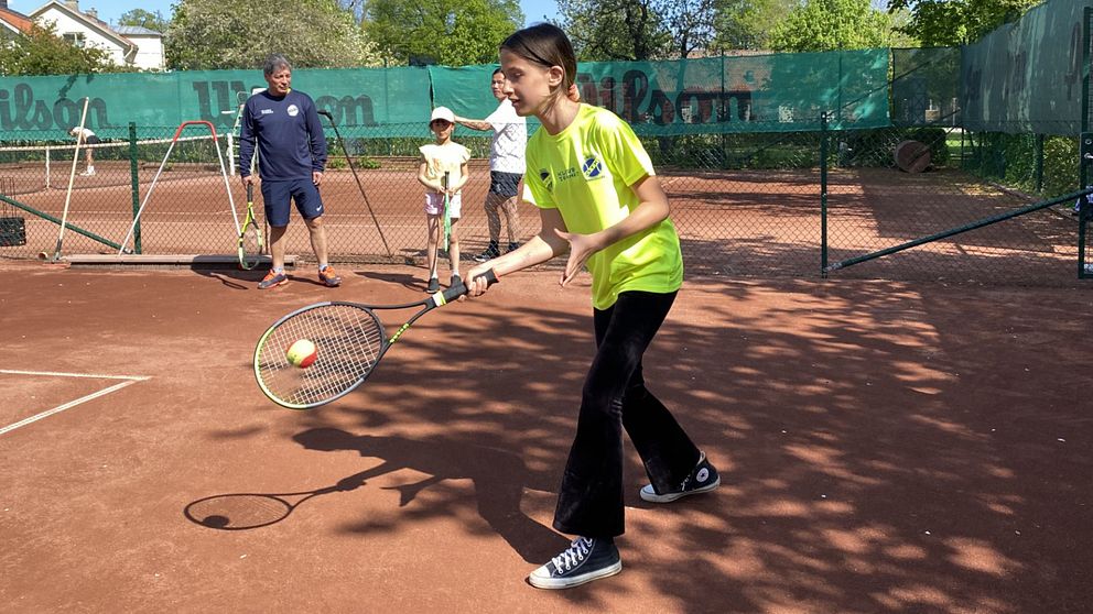 Ung tjej som spelar tennis samtidigt som några i bakgrunden tittar på.