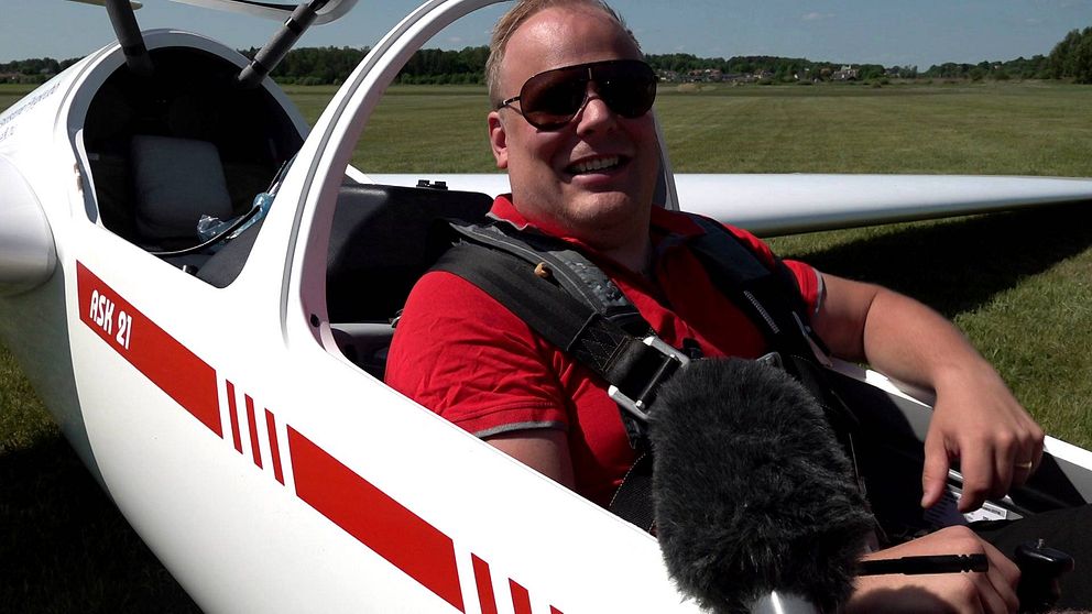Dannis Fredriksson, man med pilotglasögon ler med mikrofon framför sig i ett litet flygplan.