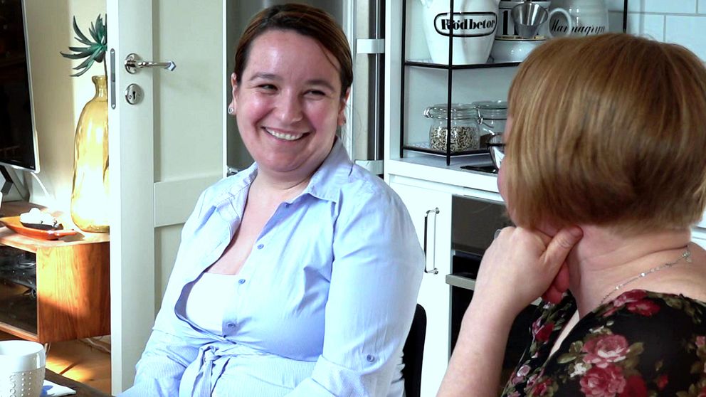 Två kvinnor sitter vid ett kökssbord. Den ena kvinnan tittar på den andra och ler. Den andra kvinnan syns i profil och tittar mot den leende kvinnan.