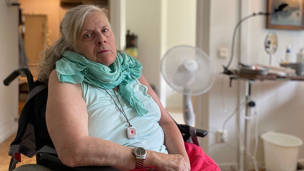 Äldre kvinna i rullstol tittar in i kameran, i bakgrunden en fläkt.
