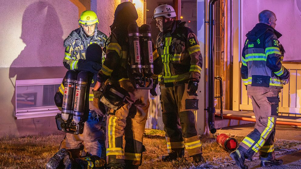 Fyra personer ur räddningstjänsten står utanför ett hus, en av dem står på knä. Två av dem har syrgasutreustning på sig.