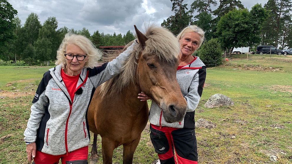 Anna-Karin och Anna står på varsin sida om en islandshäst som är mörkt brun med ljus man. På himlen bakom dem är molnen svarta och det blåser men man skymtar också solen.