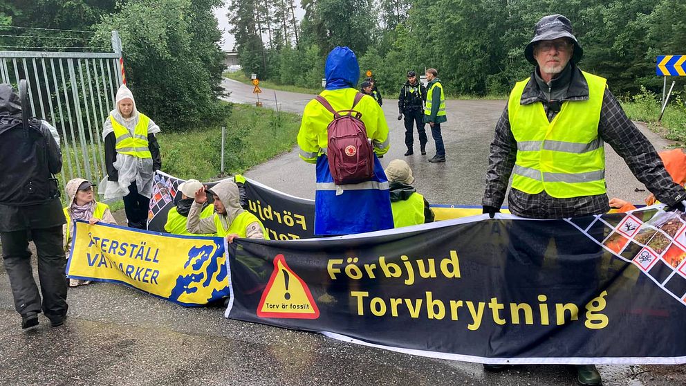 Ca sju personer i gul refelxvästar sitter på väg med banderoll märkt ”Återställ våtmarker. Torv är fossilt. Förbjud torvbrytning”. En person med Fjällräven-ryggsäck står med ryggen mot fotografen. I bakgruden står några poliser och talar med en person med reflexväst.