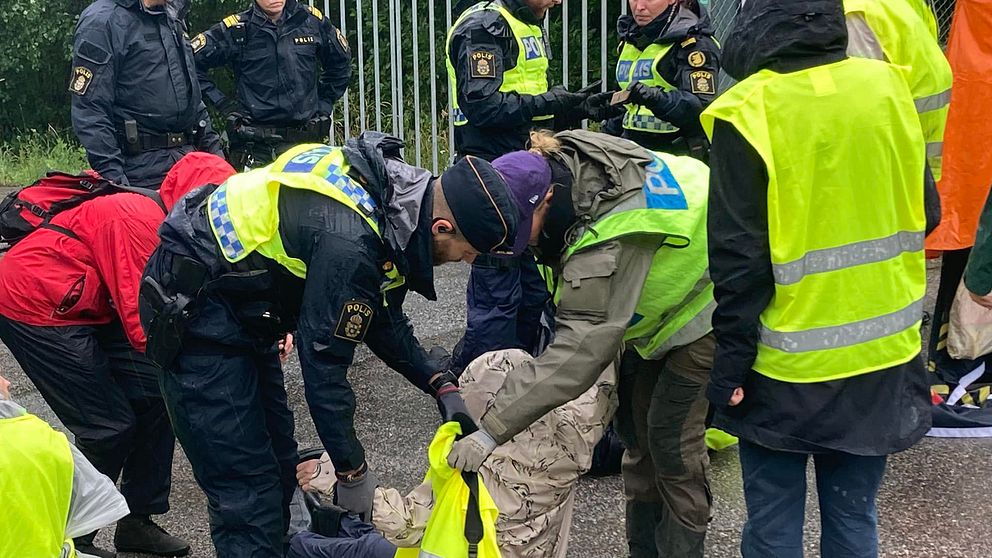 Flera poliser i gula västar försöker lyfta iväg personer som sitter på marken vid grind.