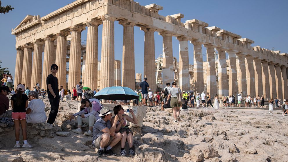 En kvinna dricker vatten i skydd från solen under ett paraply framför Parthenontemplet