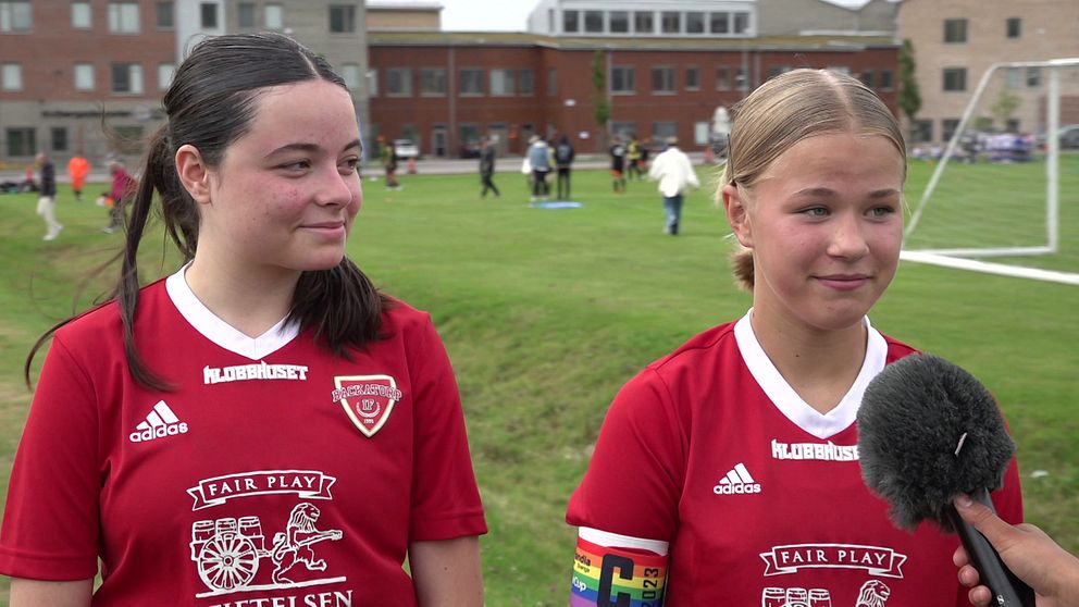 Två unga tjejer ses intervjuas, iklädda röda matchtröjor. I bakgrunden ser vi en fotbollsplan med besökare.