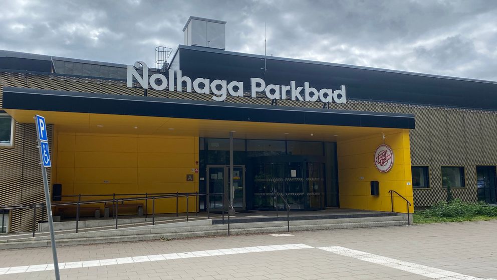 En bild på entrén till ett badhus, Nolhaga Parkbad i Alingsås. Det är en gul byggnad med badhusets namn på en skylt på taket.