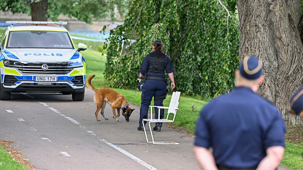 I förgrunden: En polis med händerna bakom ryggen kollar bort mot en kollega. I bakgrunden: En polis och en polishund bredvid en polisbil.