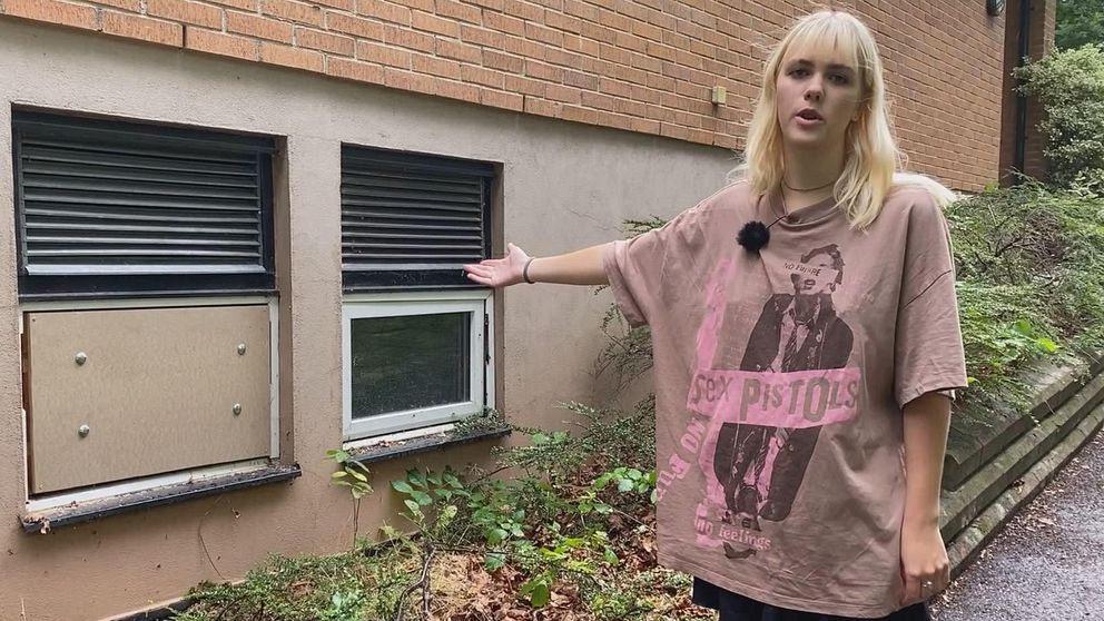 Blond kvinna tittar mot kameran och pekar mot två källarfönster varav ett är förspikat. Lila t-shirt med Sex Pistols på. Dea Nilsson, Falkvikskyrkan, Sölvesborg, Kyrkpunk.