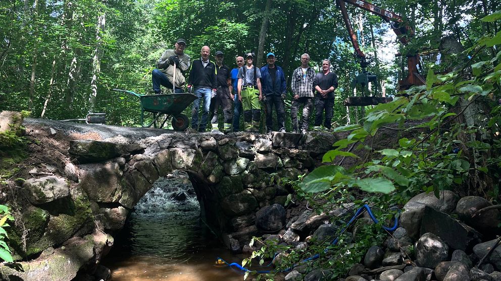 Åtta män står vid en bro, den gamla stenvalvsbron nedanför Sillegården, i skogen. En av männen har benet på en skottkärra.
