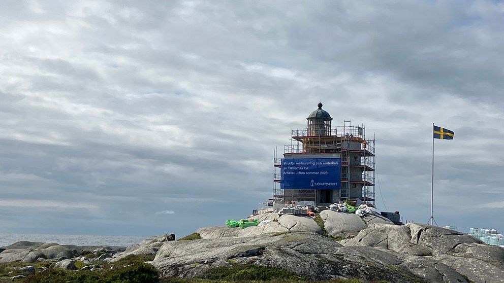 En fyr, Tistlarnas fyr, med byggnadsställning runtom står på en klippa. Bredvid fyren står en flaggstång med svenska flaggan hissad.