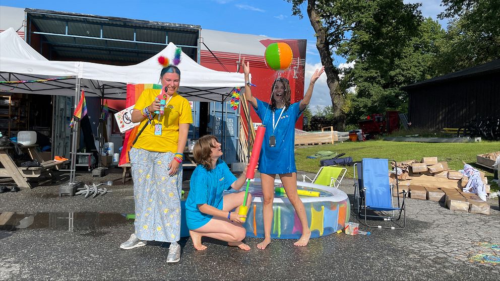 Lizzie, Nora och Stella jobbar som volontärer på Stockholm Pride och har hjälpt till att bygga upp Pride Park på Östermalms IP. På bilden sprutar volontärerna med vattenpistoler och kastar boll vid en barnpool i regnbågsfärger som är placerad vid byggboden där allt materiel och verktyg förvaras. Volontärerna skrattar.