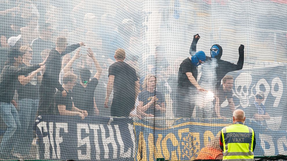 Flera maskerade supportrar står på läktaren under fotbollsderbyt mellan Östersunds FK och Gif Sundsvall i juli 2023. Runt dem virvlar rök från pyroteknik.