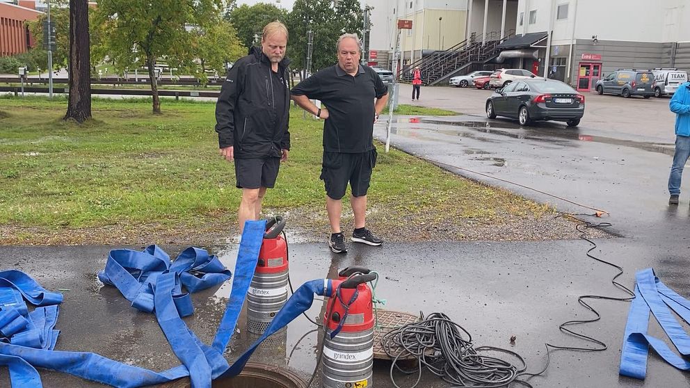 Två män står vid en brunn på en väg vid Timrå Arena och jobbar med översvämningen.