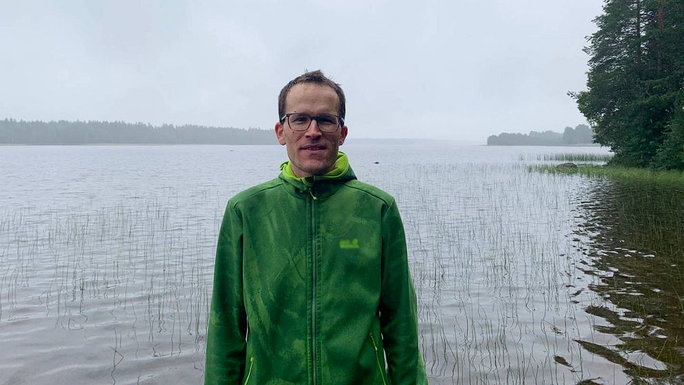 Forskaren Marcus Klaus står framför en sjö. Det är en regnig grå dag. Han har mörkt hår, grön jacka och glasögon.