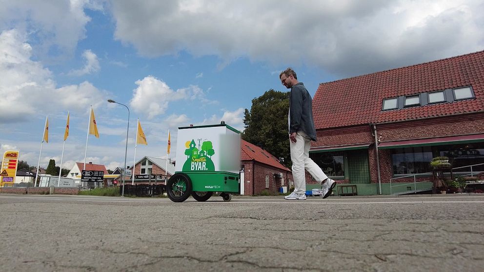 En självkörande robot formad som en låda på cykelbanan i Veberöd. På roboten står Smarta byar. Bakom roboten går produktdesigner Anton Ebel.