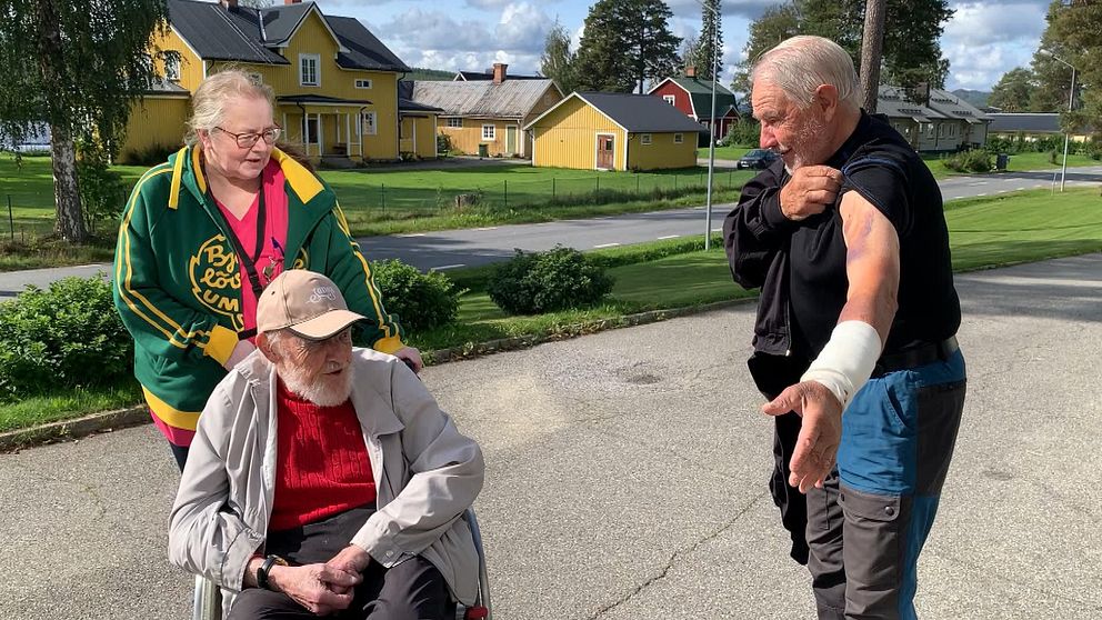 En man i rullstol och en kvinna som knuffar på tittar på en annan man som visar upp sina skador efter att ha blivit attackerad av en björn.