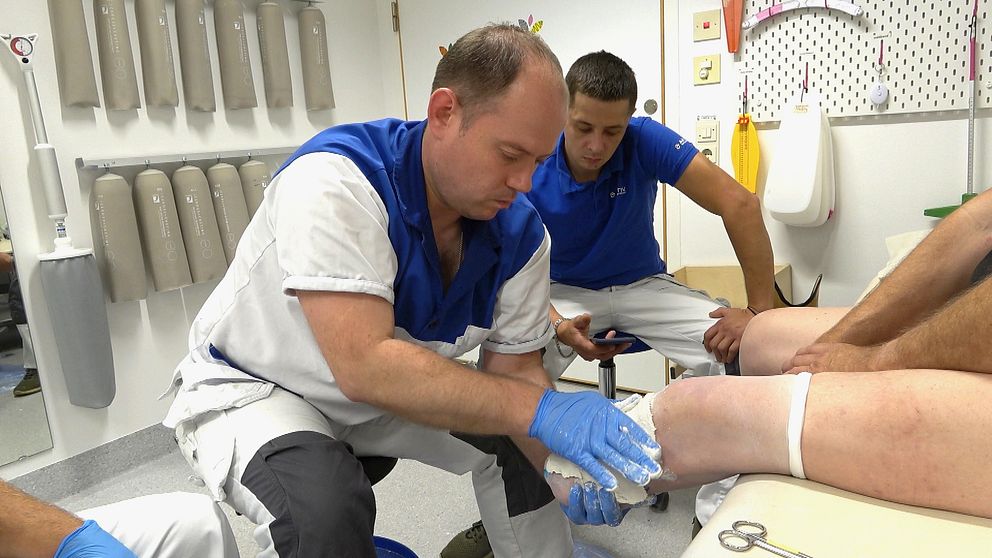 Ortopedteknikern Evhen Kaptelov från Tjernivsi i Ukraina sitter på en pall och formar gips på en patientens underben. Kollegan Vitalii Kmit sitter bredvid med en telefon i handen.