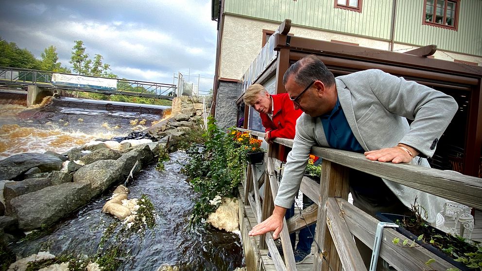 Restaurangägaren Michel Bou Antoun och Miljöpartiets språkrör Per Bolund lutar sig över räcket till Falkenberska kvarnens terrass. Michel visar med handen hur högt upp vattnet från Svartån nådde i samband med översvämningen.