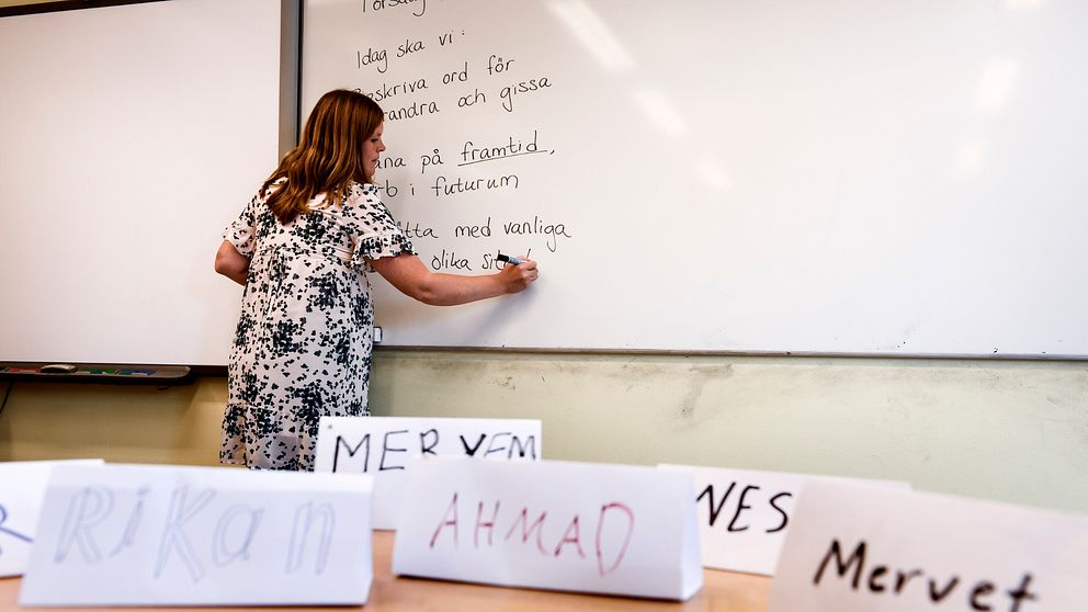 En kvinna i klänning skriver på en whiteboard. Framför henne syns namnlappar.