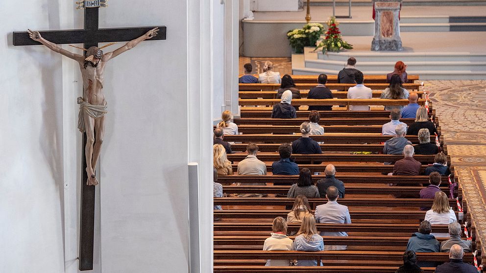 Bild inifrån en kyrka där människor sitter på kyrkbänkar.