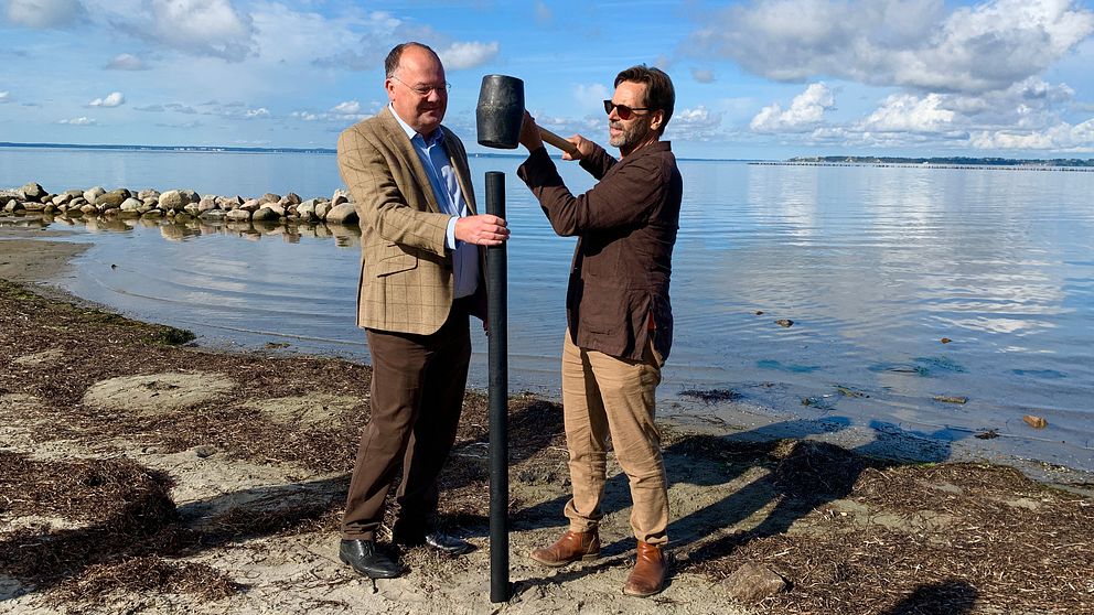 Kommunstyrelsens ordförande i Landskrona, Torkild Strandberg (L) och kallbadarföreningen Doppingarnas ordförande, Kjell Svegrup står vid havet i Landskrona. Där slår de gemensamt ned en påle i stranden som symboliserar det först ”spadtaget”.