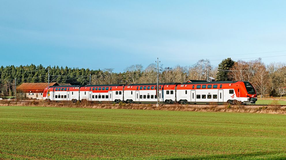 Tågmodellen ER1 ute på spåret (Tåg i Bergslagen). Dubbeldäckartåg med vit och röd målning åker genom landskap med grön åker i förgrunden och lätt blå himmel bakom.