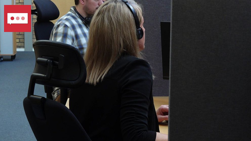 En svartklädd kvinna sitter med hörlurar framför en dator. Man ser henne snett bakifrån. Bakom henne sitter en man i rutig skjorta.