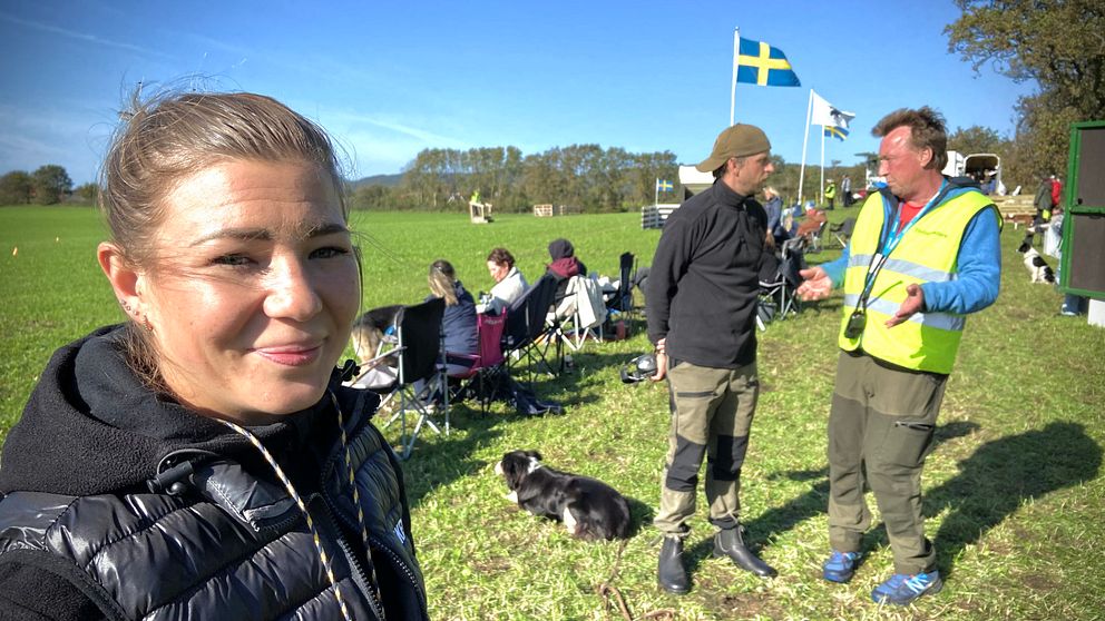 Yngre kvinna i solsken ser in i kameran. Två män till höger samspråkar.