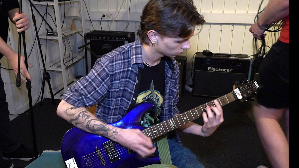 Ung man med mörkt hår och tatueringar spelar på en blå elgitarr.