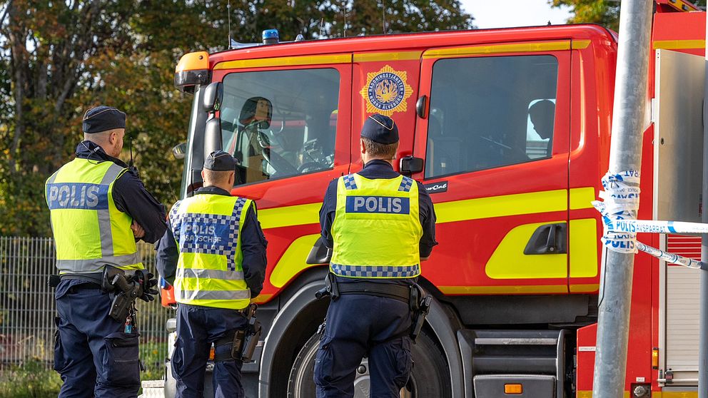 Tre poliser står med ryggen mot kameran. De har gula västar på sig. De står vid en brandbil.