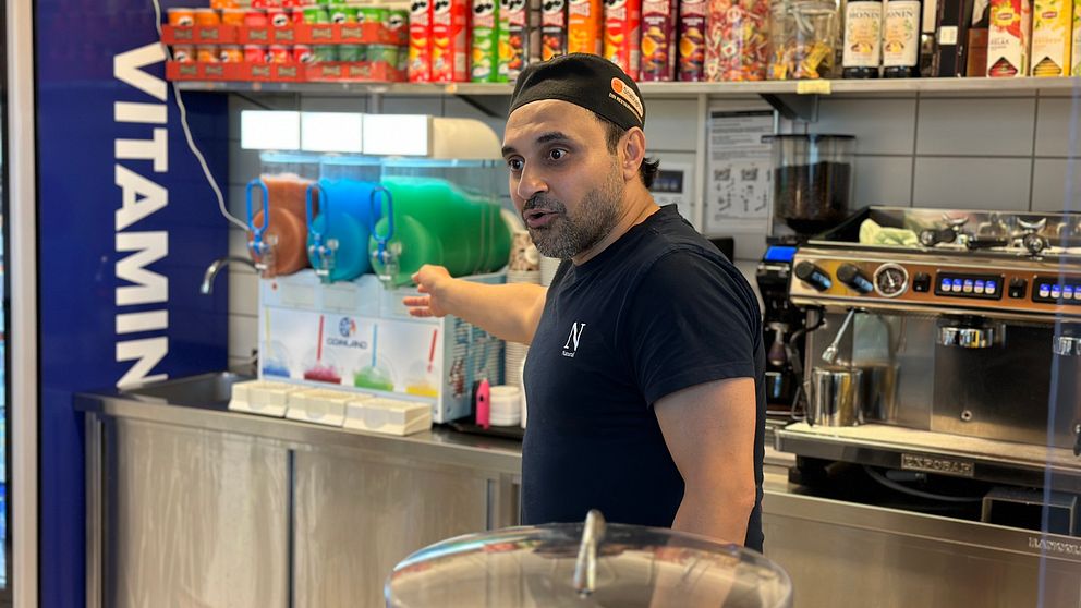 Kioskägaren Aghil Erfani Kia står vid kassan och pekar in mot kiosken, i bakgrunden syns olika sorters chips och en slushimaskin.