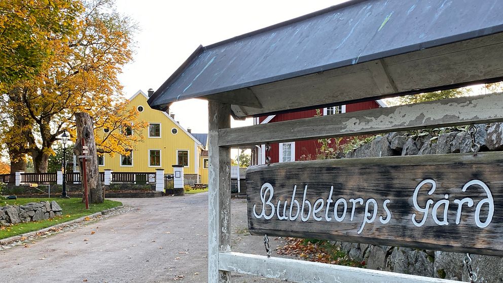 Skylt på Bubbetorps gård med vit text, i bakgrunden ett rött och ett gult hus samt ett höstfärgat träd.