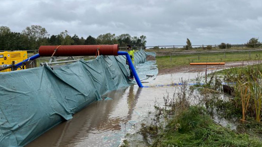 Pressenningstäckt översvämningsskydd i form av en vall, vatten i förgrunden och en grävmaskin i bakgrunden