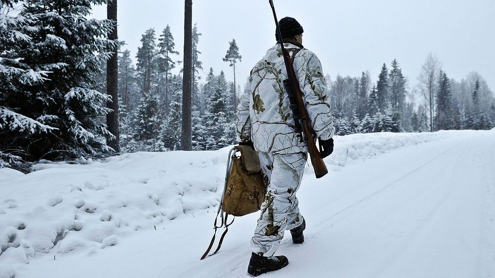 En jägare i vinterlandskap iklädd vita kamouflagekläder med bössan på axel.