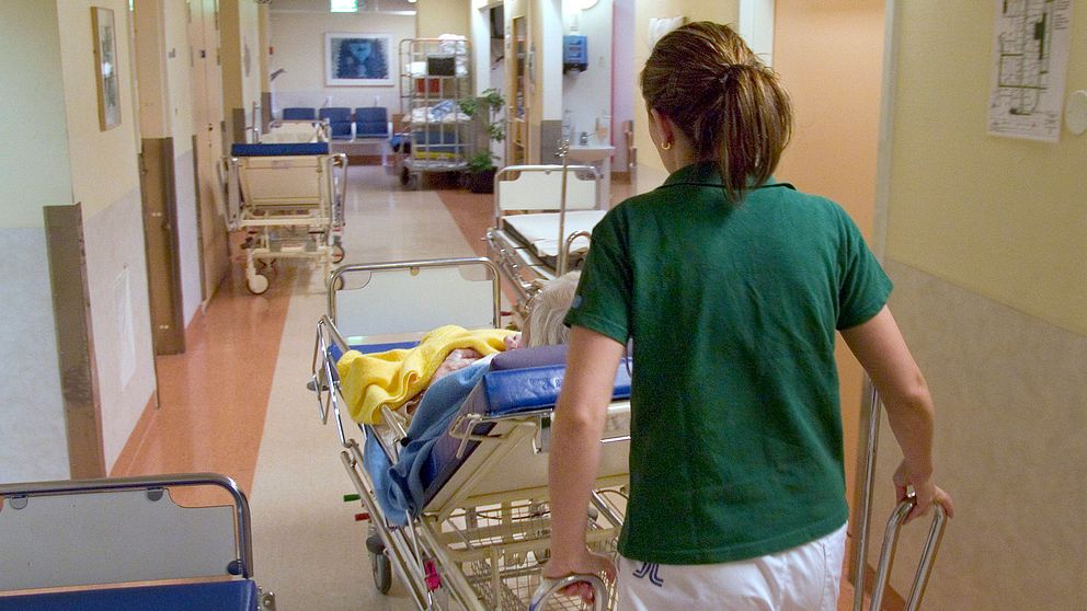 Sjukvårdspersonal flyttar patient i sjukhussäng i korridor.