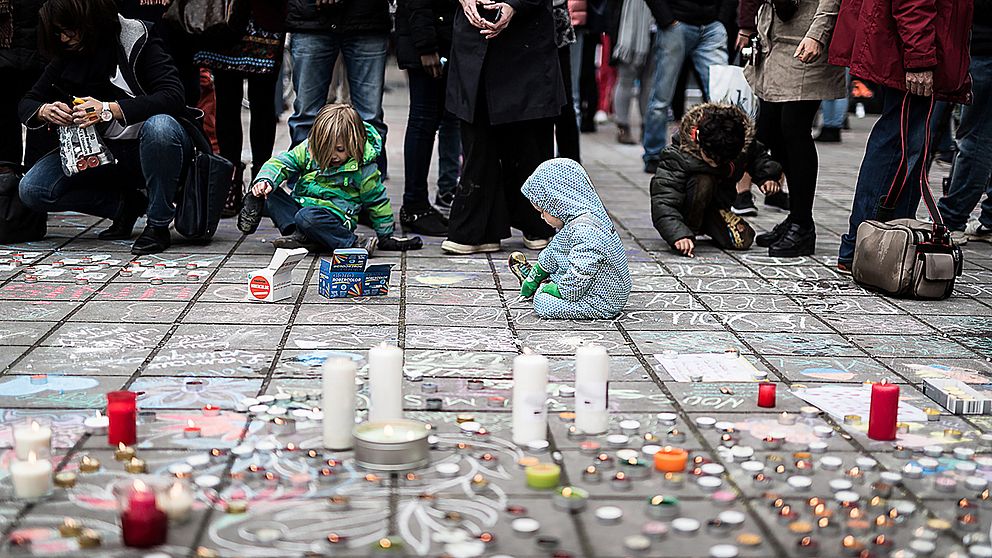 Några små barn sitter på marken vid en minnesplats för terrordådens offer i Bryssel, omgivna av sörjande människor.