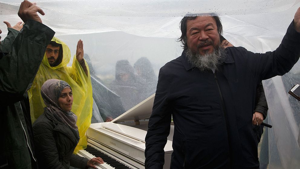 Ai Weiwei håller upp en presenning så att den syriska pianisten Nour al Khzam ska kunna spela i flyktinglägret Idomeni.