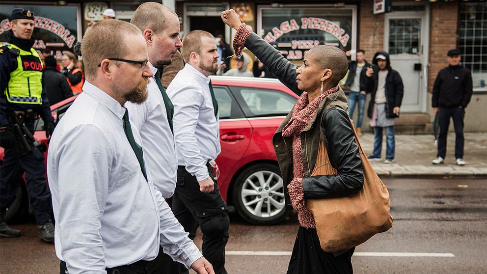En bild på en kvinna, med en höjd knuten näve, som står i vägen för tre demonstrerande män från Nordiska motståndsrörelsen.