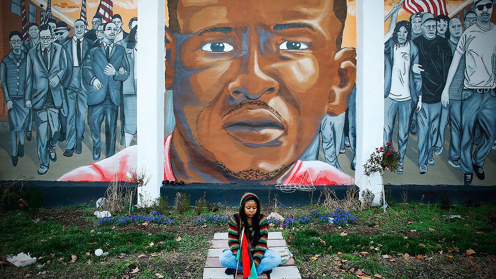 En kvinna sitter framför en väggmålning som föreställer Freddie Gray och som satts upp intill platsen där han greps.